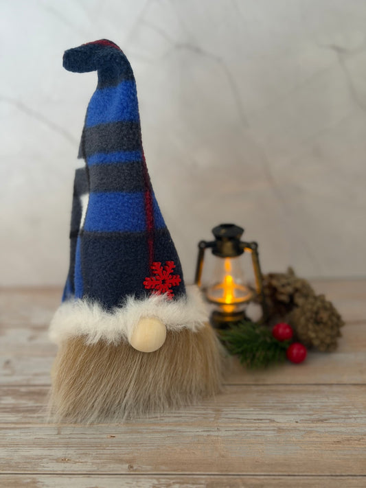 A festive Nordic style gnome decoration with a tall blue and navy striped fleece hat, fluffy beige beard, white faux-fur trim, and a small red snowflake ornament, displayed on a wooden surface beside a lantern and holiday decor. Handmade by Northern Nomes.