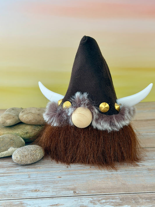 A decorative Viking-style gonk figurine with a tall dark brown hat, white horn-like side pieces, brown beard, faux fur trim, gold studs, and a round wooden nose, displayed on a weathered wood surface with stones and a soft pastel background.