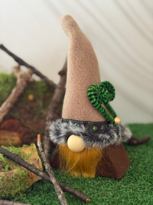 A small gnome decoration with a tall tan hat, green leaf accent, gray faux-fur trim, ginger beard, and brown base, displayed on artificial grass with branches and moss. Handmade by Northern Nomes.