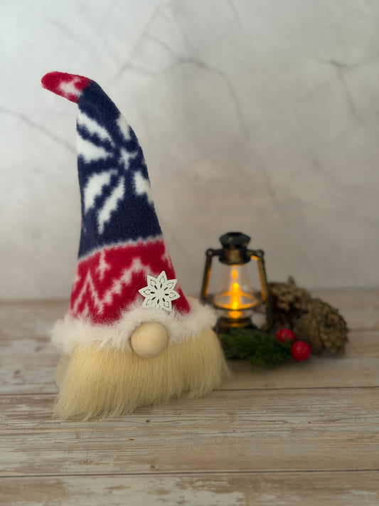 A small Christmas Nordic gnome decoration with a tall red, white, and navy patterned hat, a fluffy cream beard, and a wooden nose, styled on a rustic wooden surface with a lantern, pinecones, and berries in the background.