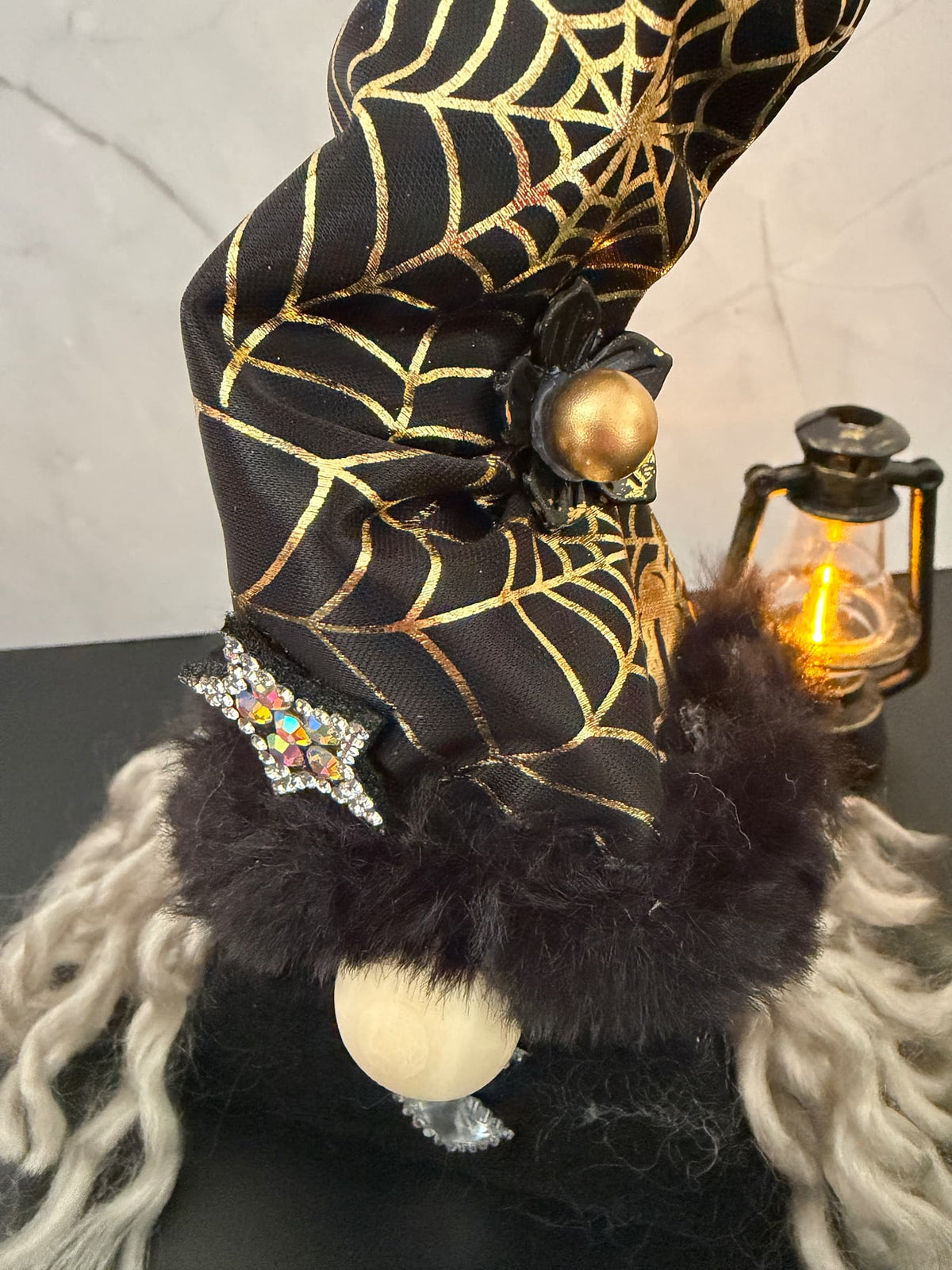 Close up of a Halloween witch gnome hat decoration in black and gold spiderweb patterns, a black and gold accent, a jewelled star embellishment, dark faux fur trim, and long white curly hair, displayed on a dark surface with a lantern in the background.