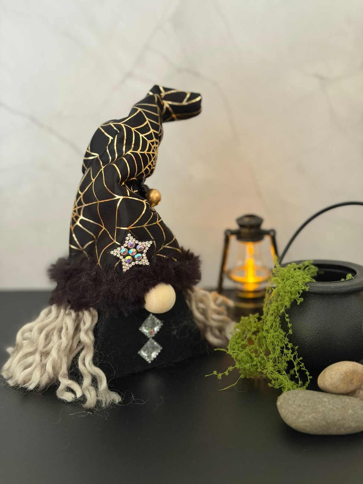 A Halloween witch gnome ornament with a black hat decorated with gold spiderwebs, a black faux-fur trim white curly hair, and rhinestone accents, styled on a dark tabletop next to a cauldron, moss, stones, and a lantern.
Handmade by Northern Nomes.
