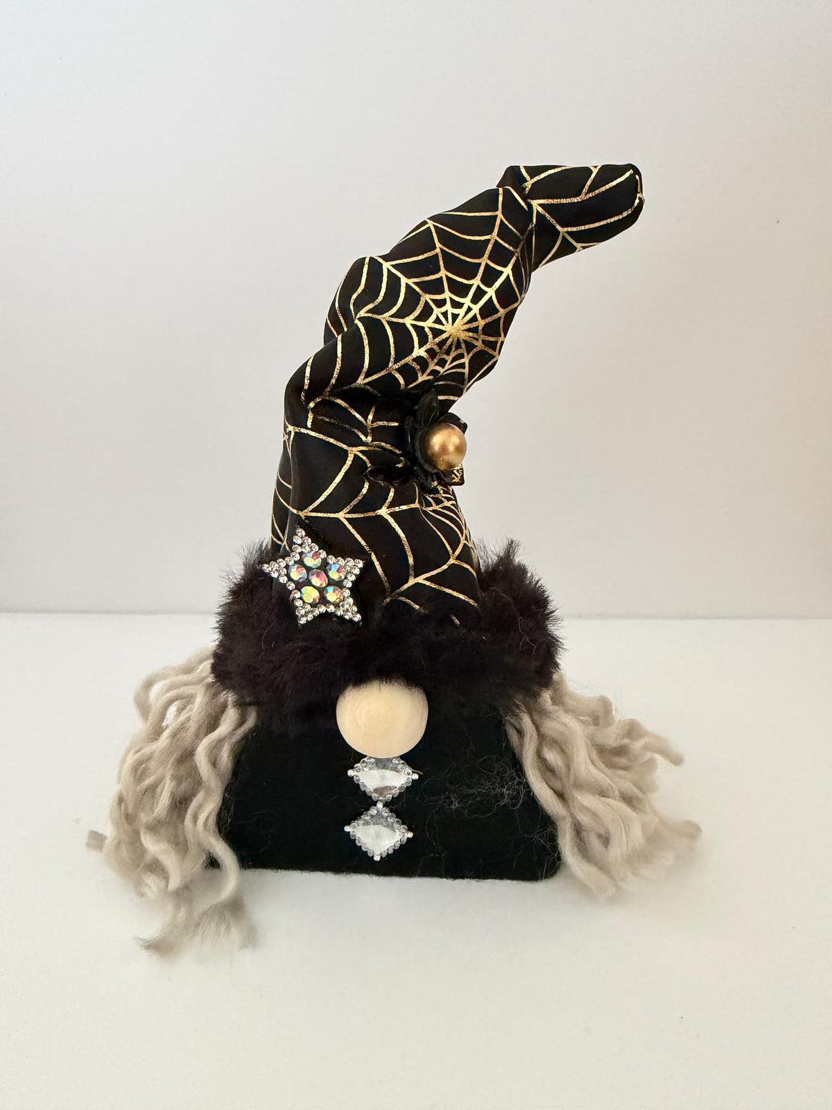 A handmade Halloween witch gnome ornament with a black hat decorated with gold spiderwebs, a black faux-fur trim white curly hair, and rhinestone accents, styled on a white background. 
Path of the Hidden Folk Gonk collection by Northern Nomes.