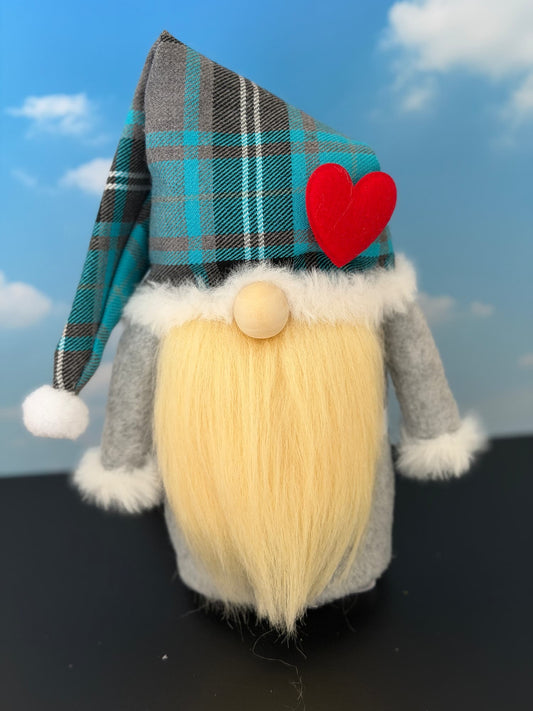 A handmade gnome character with a long blonde faux-fur beard, gray felt body, white faux-fur trim, a plaid blue, gray, and black nightcap styled hat, and a red felt heart on the side, standing against a blue sky backdrop. Made by Northern Nomes.