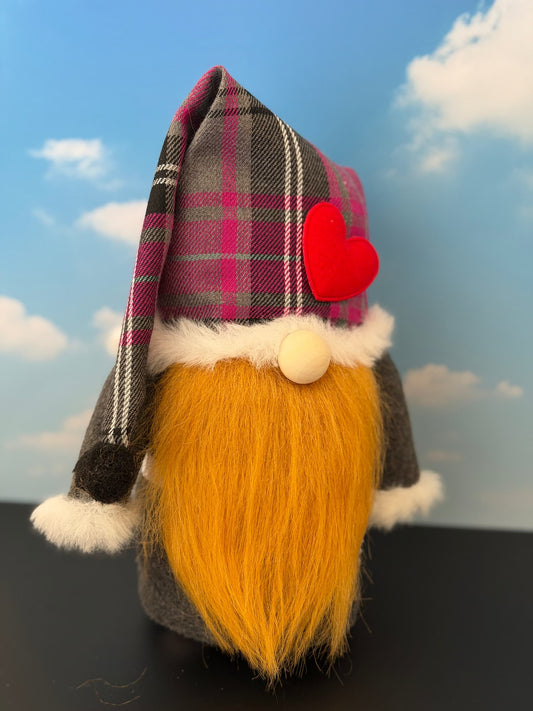 A decorative gnome figurine with a bright ginger beard, gray body, white faux-fur trim, and a plaid hat in pink, gray, and black with a red heart accent, shown against a blue sky backdrop. Handmade character from Northern Nomes.
