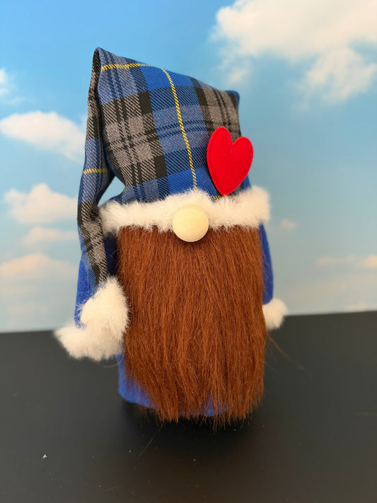 A small decorative gnome figurine with a blue plaid nightcap style hat, white faux fur trim, a red heart accent, and a long brown faux-fur beard, standing on a dark surface against a blue sky backdrop with clouds. Handmade character gnome by Northern Nomes.