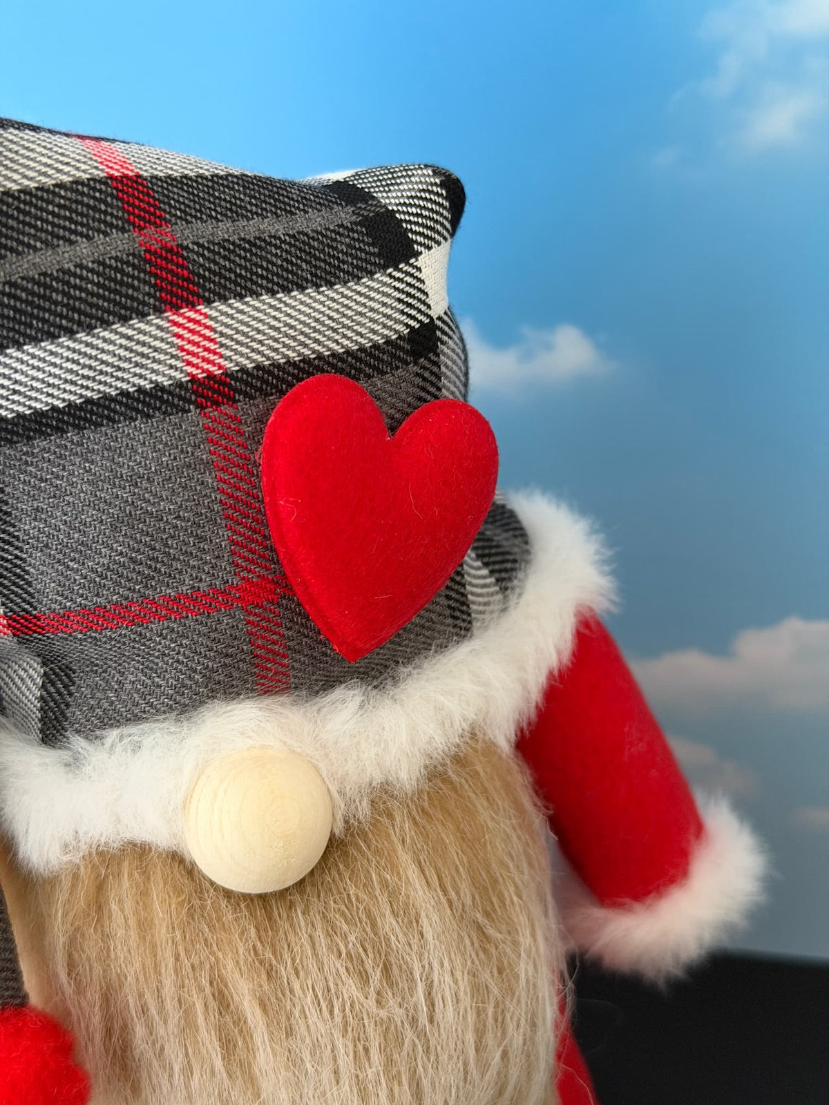 A close-up of a gnome character wearing a black, white, and red plaid hat with a red felt heart and white faux fur trim, against a blue sky background. Handmade by Northern Nomes.