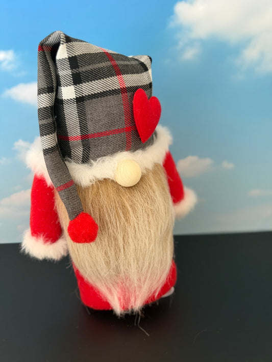 A handmade gnome figurine by Northern Nomes from the Scottish Dreamers collection, wearing a gray and black plaid hat with red trim, a red body with white faux fur trim, a beige faux-fur beard, and a red felt heart on the hat, set against a blue sky backdrop.