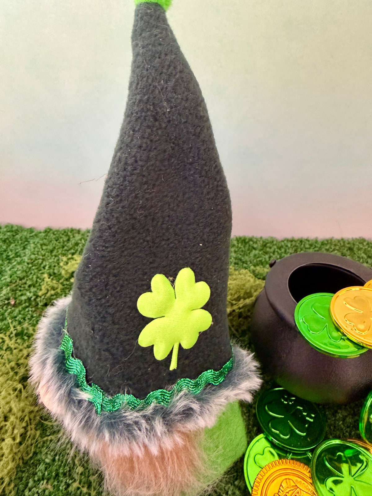 A St. Patrick's Day gnome decoration featuring a tall dark green felt hat with a bright green shamrock, green trim, and faux fur trim, displayed on artificial green grass next to a small black pot of shiny green and gold coins.