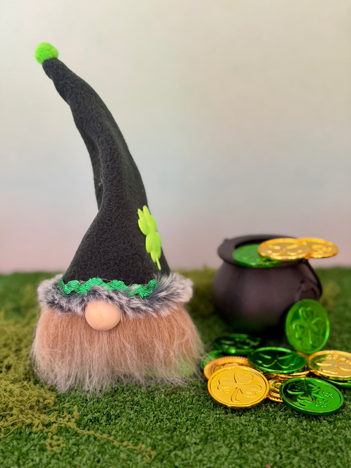 A handmade St. Patrick's Day lucky Leprechaun gnome figurine with a tall dark green hat, green shamrock accent, green trim, and a beige beard, posed next to a black pot and shiny gold and green coins on artificial green grass against a soft pastel background. Made by Northern Nomes.
