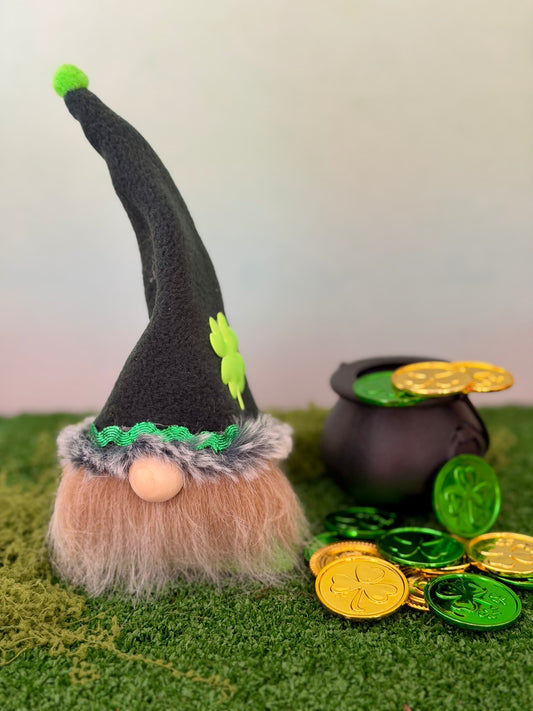 A handmade St. Patrick's Day lucky Leprechaun gnome figurine with a tall dark green hat, green shamrock accent, green trim, and a beige beard, posed next to a black pot and shiny gold and green coins on artificial green grass against a soft pastel background. Made by Northern Nomes.