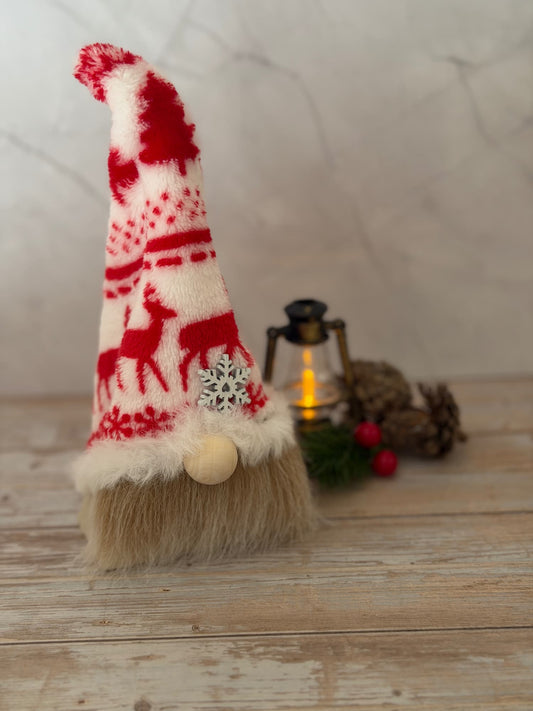 A festive Christmas gnome decoration with a red and cream knit-patterned hat featuring reindeer motifs, a fluffy beige beard, and a small wooden nose, styled on a rustic wooden surface with a lantern and pinecones in the background. From the Northlight Folk a Nordic style gonk handmade by Northern Nomes.