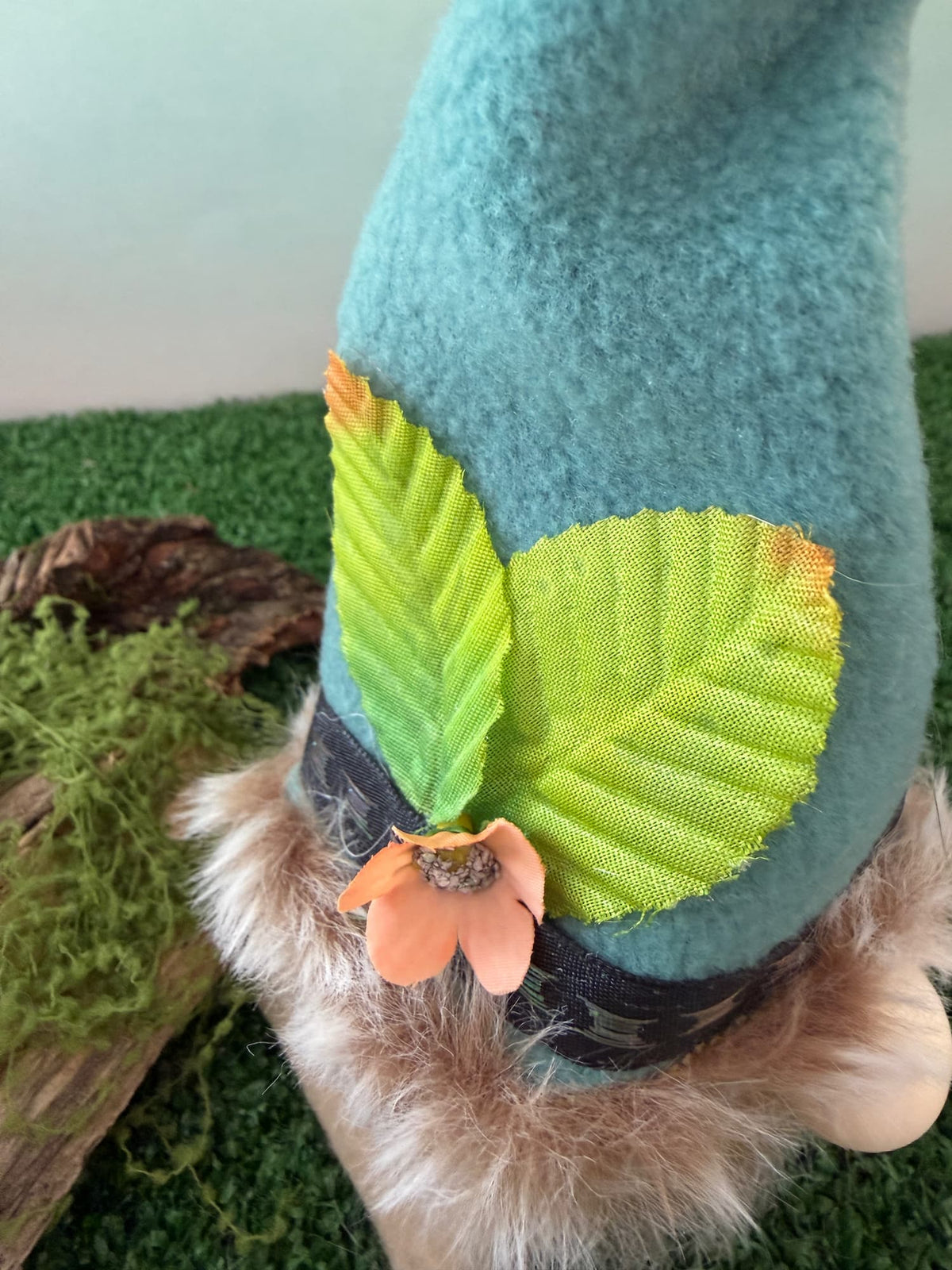 Close up of a handmade gnome figurine with a tall light greenish hat decorated with green leaves and an orange flower, set on artificial grass with a mossy log prop.