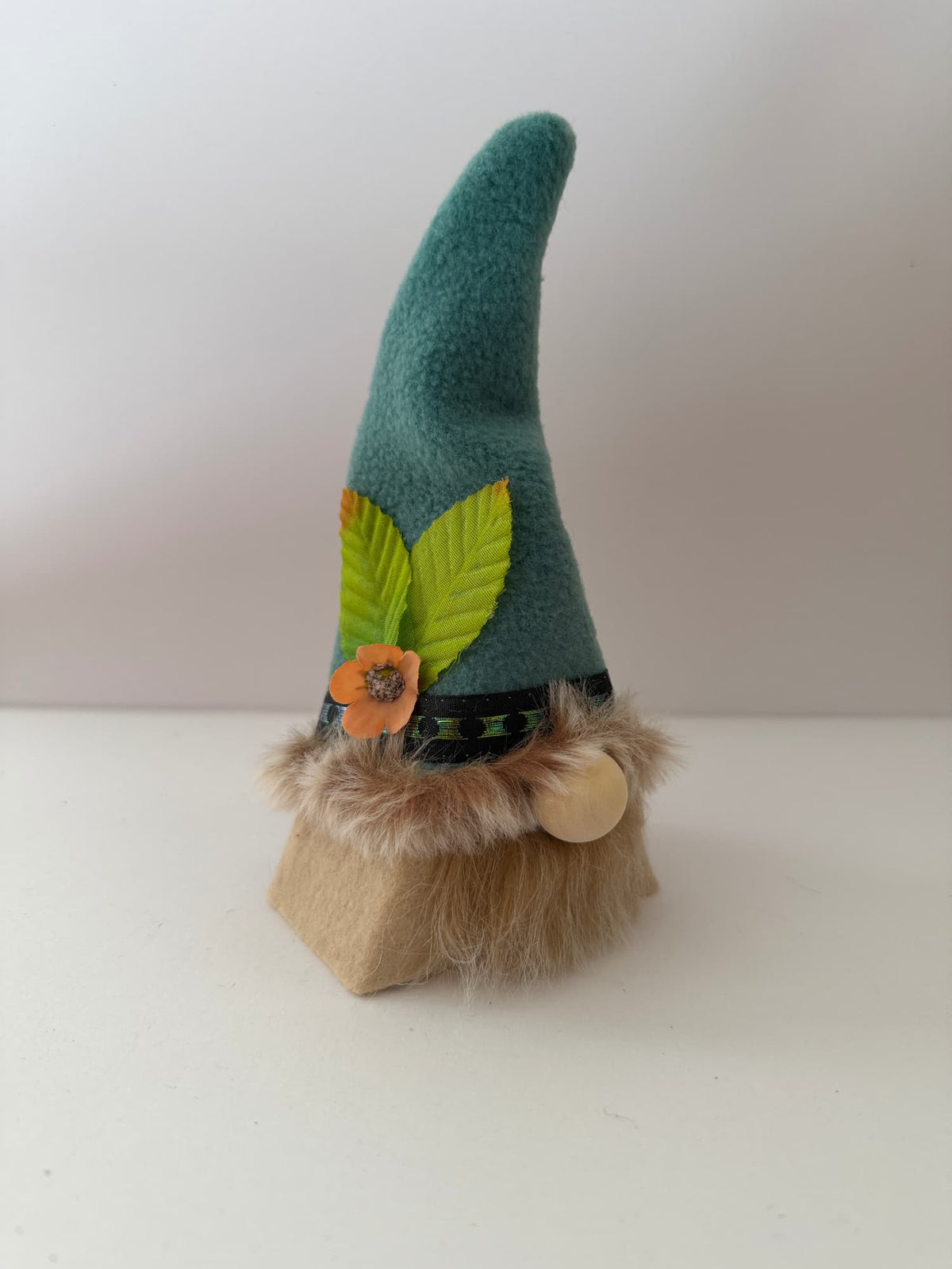 A small felt gnome figurine with a tall teal hat, green leaf accents, an orange flower, a tan beard, and a round wooden nose, standing on a plain light background. Handmade by Northern Nomes.