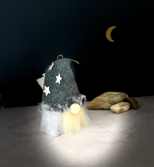 A small gnome ornament with a dark grey fleece hat decorated with white stars, a fluffy cream beard, and a light gray body, standing on a tabletop against a dark background with a crescent moon and stones. Handmade by Northern Nomes.