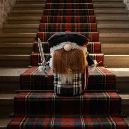 A Scottish-themed decorative gnome figurine with a long brown beard, black and white plaid kilt, and black bonnet sits on a set of stone stairs with red tartan carpeting. Handmade by Northern Nomes.