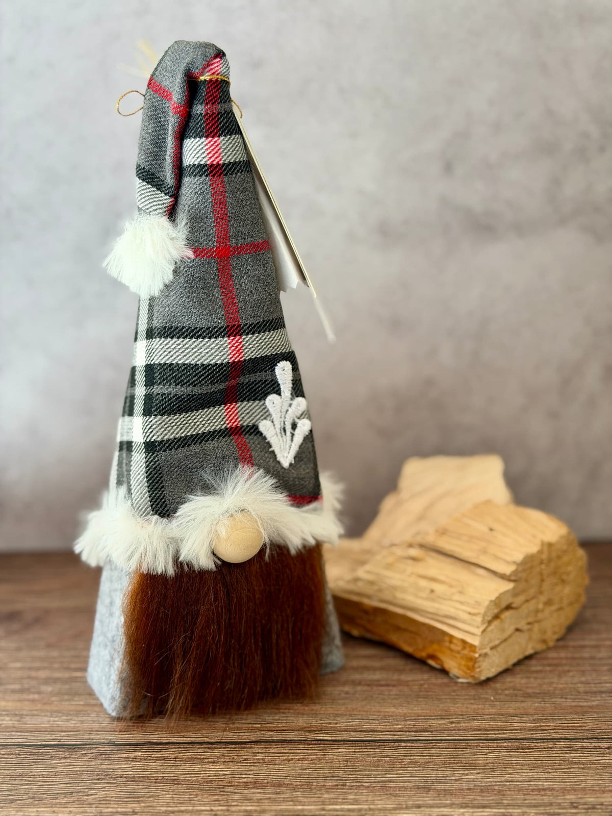 A decorative gnome figurine wearing a gray, black, and red plaid pointed hat with white faux fur trim and a brown faux-hair beard, standing on a wooden surface next to a small piece of wood against a neutral gray background. Handmade using reclaimed timber by Northern Nomes.
