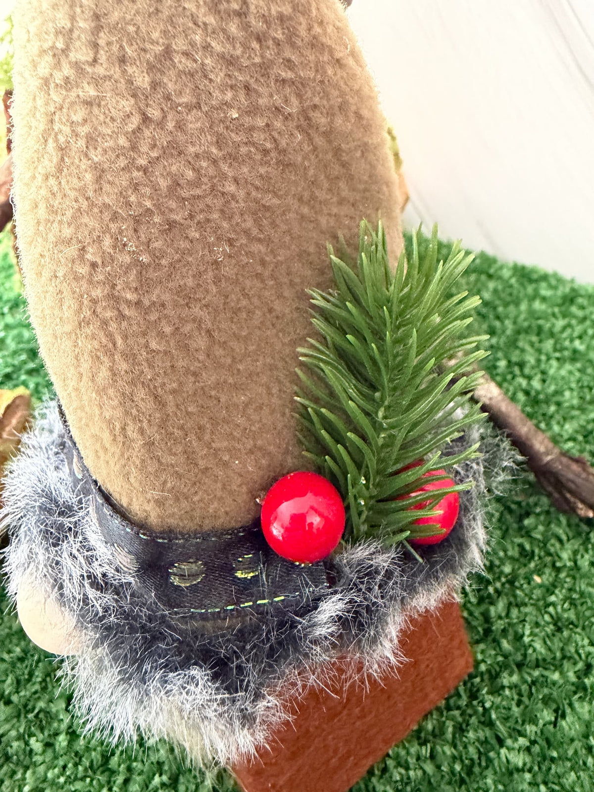 A close-up of a decorative gnome figure lfeaturing a tan textured fleece hat, a black trim with gray faux fur trim, two red berries, and a green pine sprig, set against artificial green grass.