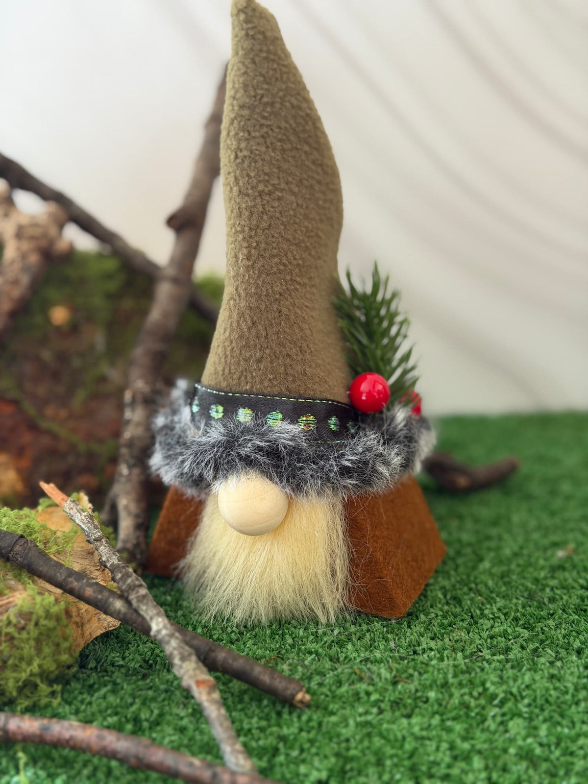 A small gnome figurine with a tall greenish felt hat, fluffy cream beard, gray faux-fur trim, and a red berry accent, posed on green artificial turf with branches and moss in the background. Handmade by Northern Nomes.