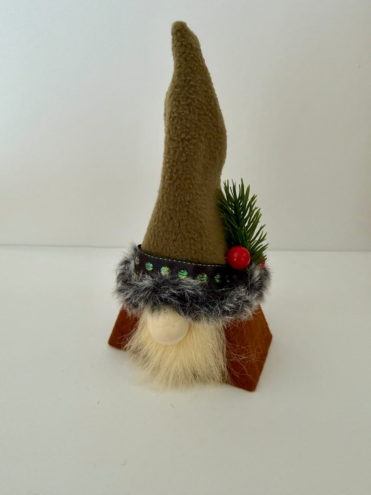 A small gnome figurine with a tall greenish fleece hat, gray faux fur trim, green pine needles, a red berry, and a cream beard, standing on a brown base against a plain white background. Made by Northern Nomes.