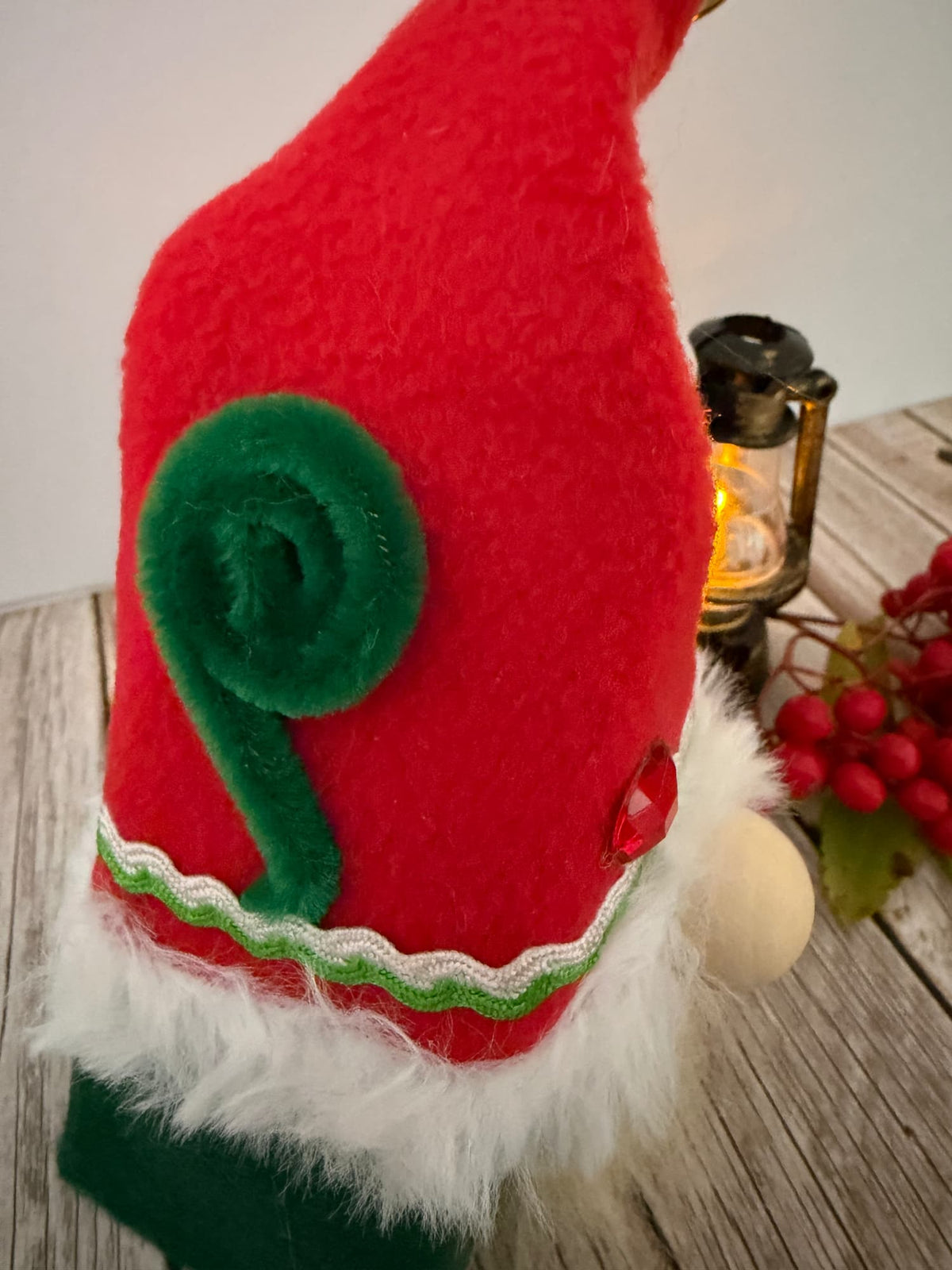 A close-up of a red and green Christmas Elf gnome hat with a green spiral accent, white faux-fur trim, and a small red button, shown on a rustic wooden surface with a lantern and berries in the background.