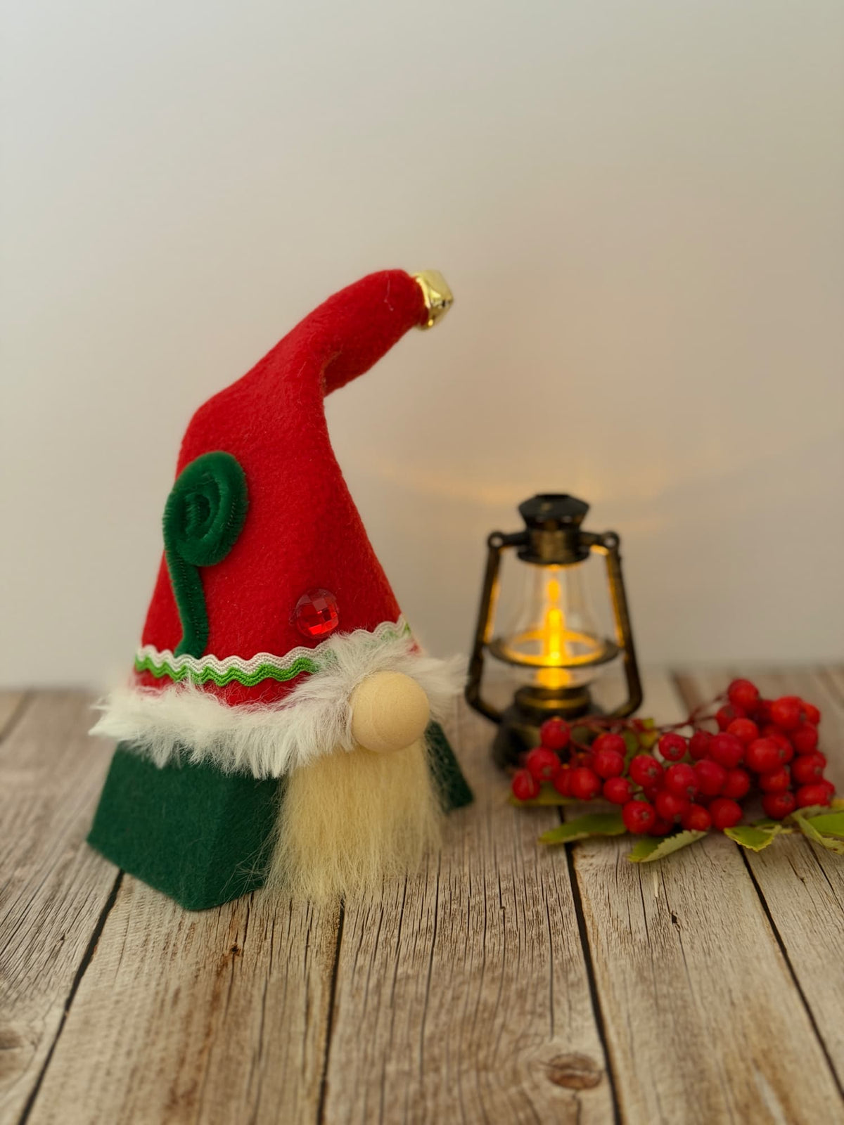A small handmade Christmas Elf gnome decoration with a red hat, green base, white faux fur trim, and cream beard, styled on a rustic wooden surface beside a lit lantern and red berries against a plain light background. From the Festive Folk collection by Northern Nomes.
