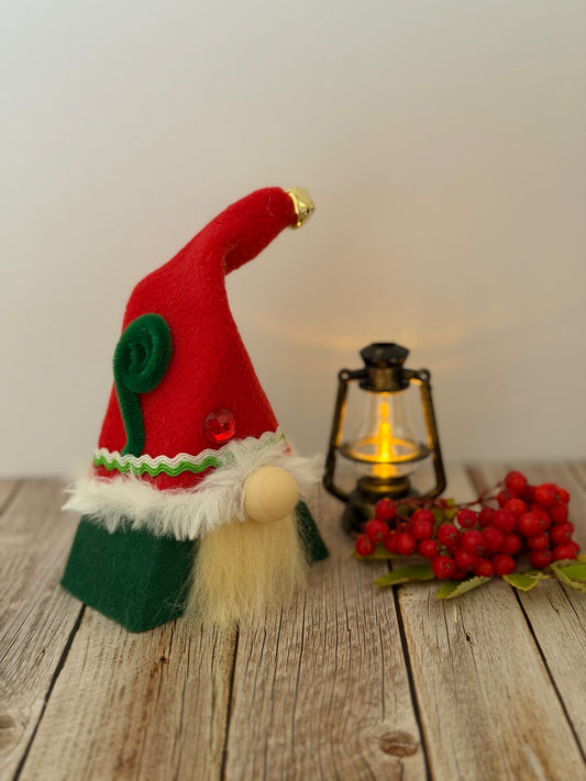 A small handmade Christmas Elf gnome decoration with a red hat, green base, white faux fur trim, and cream beard, styled on a rustic wooden surface beside a lit lantern and red berries against a plain light background. From the Festive Folk collection by Northern Nomes.