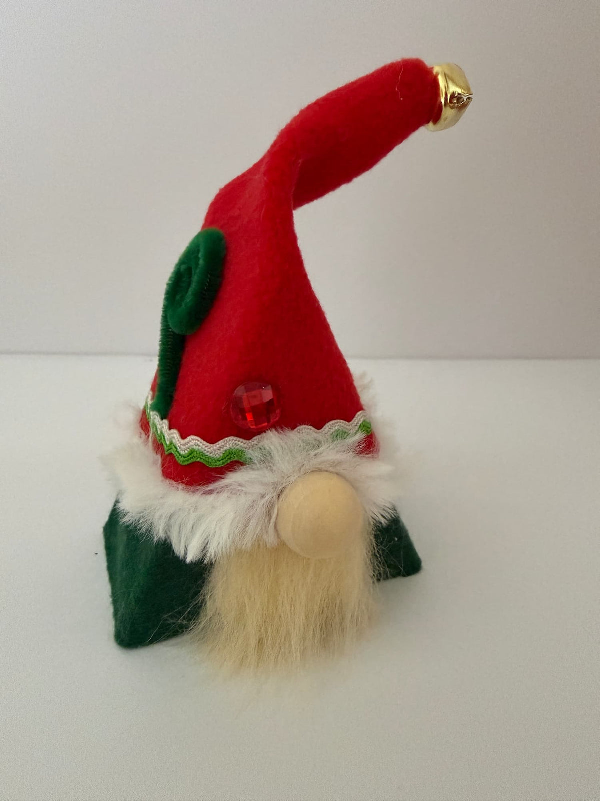 A small felt Christmas Elf gnome decoration with a red pointed hat tipped with a gold jingle bell, green side panels, a white faux-fur trim, a red jewel accent, and a beige beard, photographed on a plain light background. Handmade by Northern Nomes.