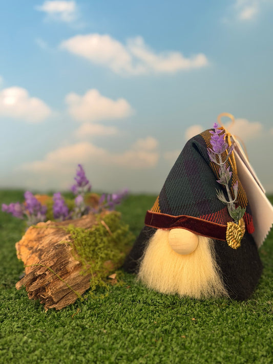 A small gnome figurine with a cream beard and black hat sits on artificial grass beside a log, with a blue sky backdrop and purple heather flowers in the background. Handmade by Northern Nomes.