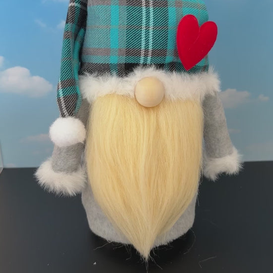 Video of a Northern Nomes Scottish Dreamer character gnome with a turquoise blue and gray plaid hat, white faux-fur trim, a red heart, with small nose peeking out from underneath the hat and a long blonde faux-hair beard, standing on a dark surface against a blue sky backdrop with clouds.