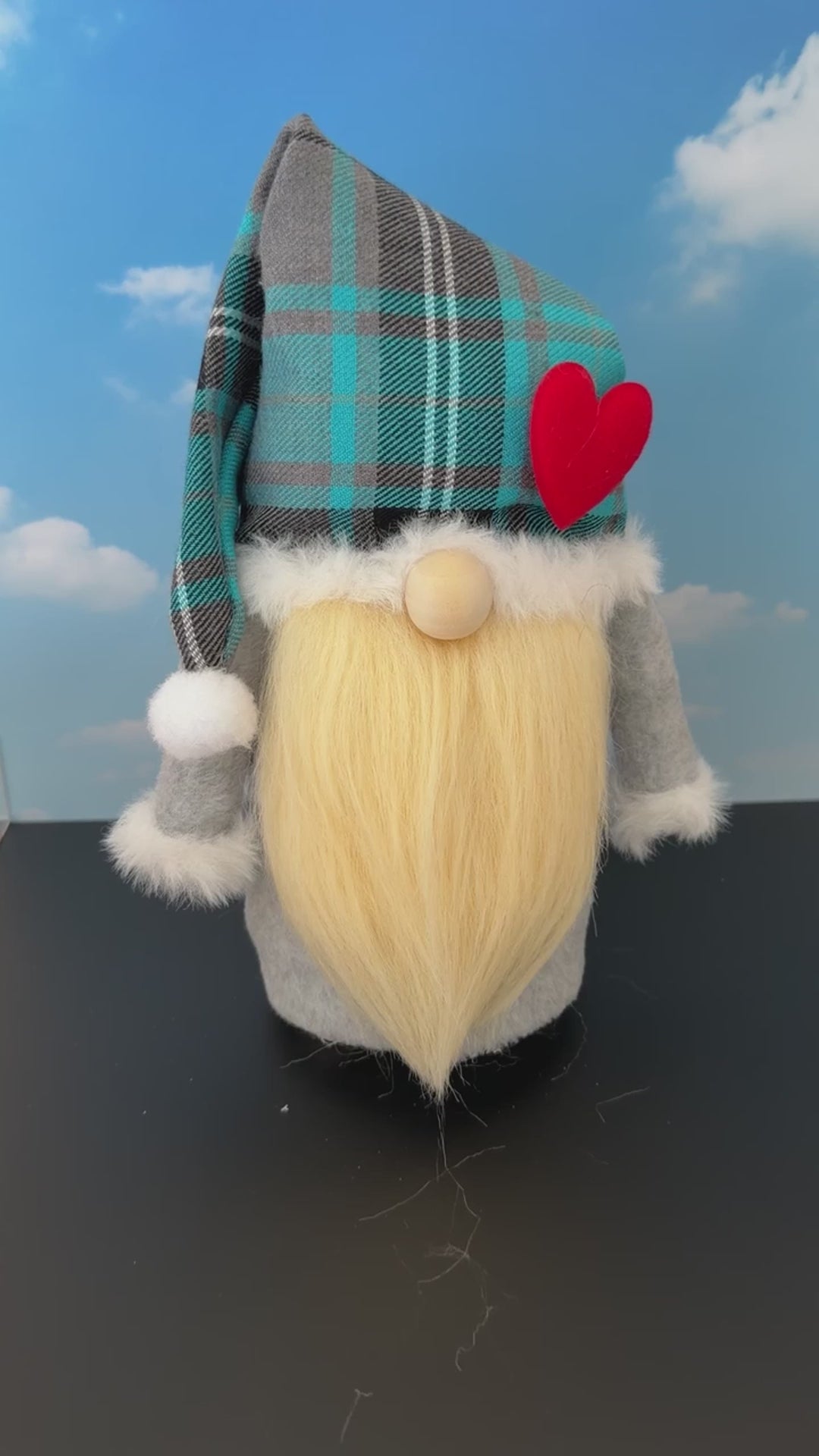 Video of a Northern Nomes Scottish Dreamer character gnome with a turquoise blue and gray plaid hat, white faux-fur trim, a red heart, with small nose peeking out from underneath the hat and a long blonde faux-hair beard, standing on a dark surface against a blue sky backdrop with clouds.