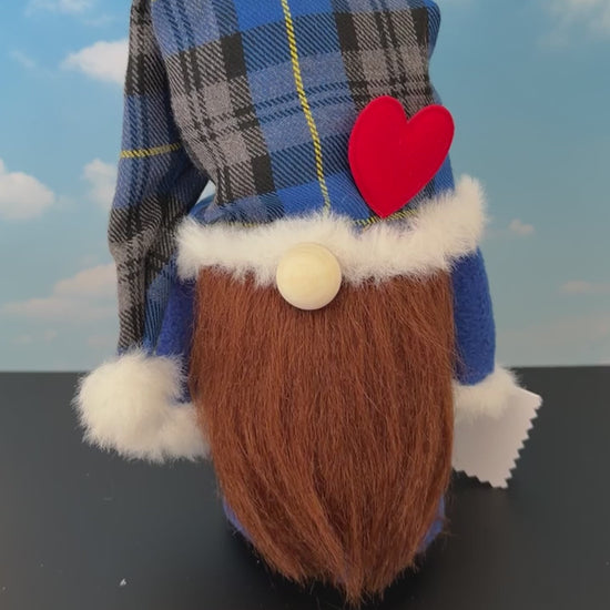 Video of a Northern Nomes character gnome figurine with a blue and gray plaid hat, white faux-fur trim, a red heart-shaped nose, and a long brown faux-hair beard, standing on a dark surface against a blue sky backdrop with clouds.