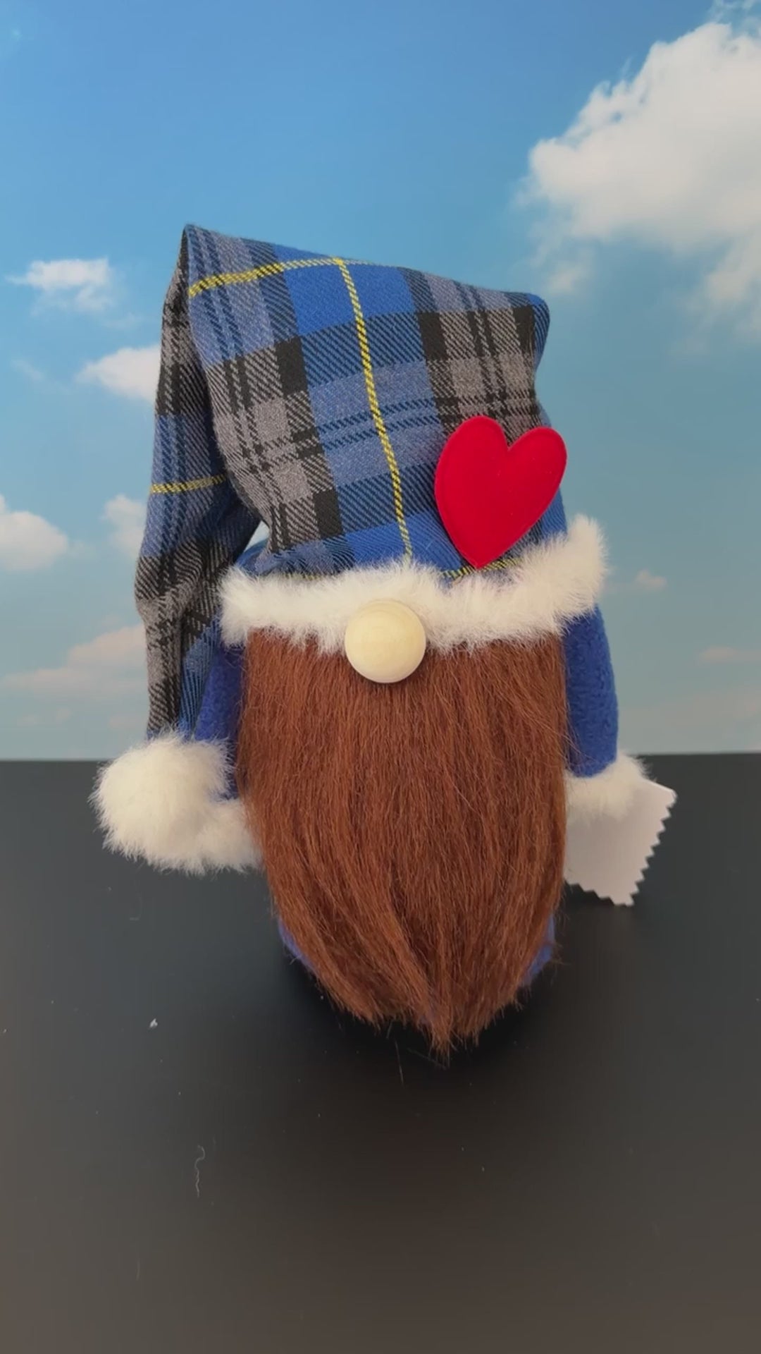 Video of a Northern Nomes character gnome figurine with a blue and gray plaid hat, white faux-fur trim, a red heart-shaped nose, and a long brown faux-hair beard, standing on a dark surface against a blue sky backdrop with clouds.