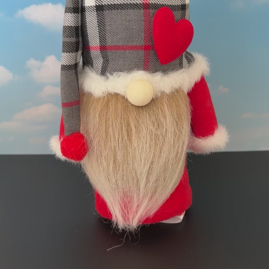 Video of a Northern Nomes character gnome figurine with a red, green and black tartan plaid hat, white faux-fur trim, a red heart, a wooden nose, and a long fawn faux-hair beard, standing on a dark surface against a blue sky backdrop with clouds.