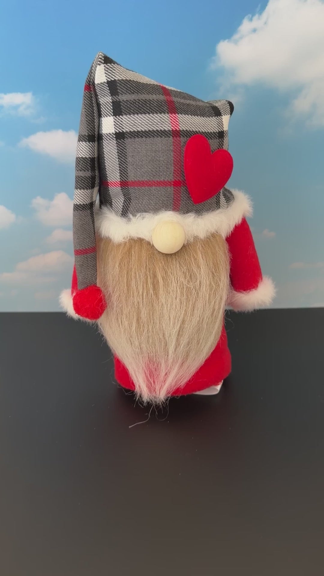 Video of a Northern Nomes character gnome figurine with a red, green and black tartan plaid hat, white faux-fur trim, a red heart, a wooden nose, and a long fawn faux-hair beard, standing on a dark surface against a blue sky backdrop with clouds.