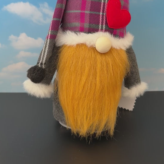 Video of a Northern Nomes Scottish Character gnome featuring a purple and grey plaid hat, white faux-fur trim, a red heart-shaped nose, and a long brownish faux-hair beard, standing on a dark surface against a blue sky backdrop with clouds.