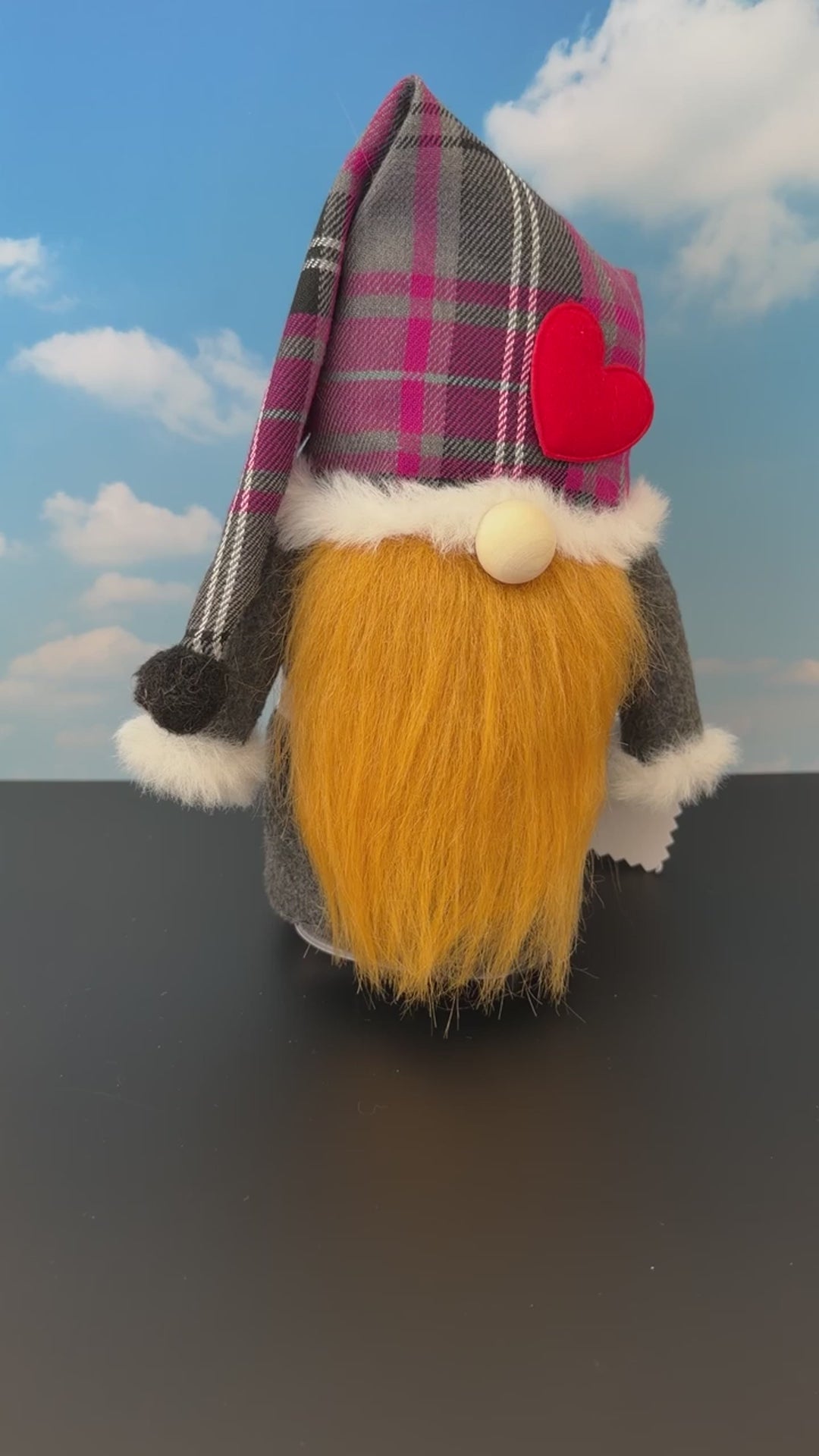 Video of a Northern Nomes Scottish Character gnome featuring a purple and grey plaid hat, white faux-fur trim, a red heart-shaped nose, and a long brownish faux-hair beard, standing on a dark surface against a blue sky backdrop with clouds.